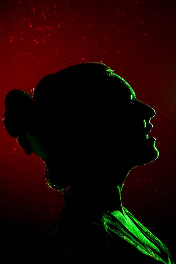 A portrait shaped image of a woman, partially silhouetted against a red background. a green gelld light to the woman's right provides a rim of light to her face and throat.