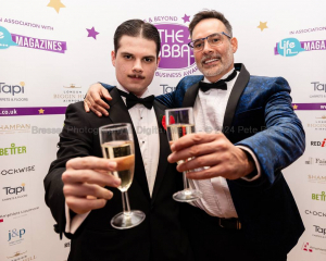 Two smartly dressed men with filled champagne flutes outstretched in front of them. Behind is a logo backdrop