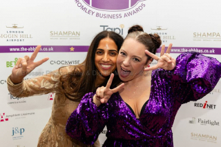 Two smartly dressed women posed with hands showing peace signs with their fingers, in front of them. Behind is a logo backdrop