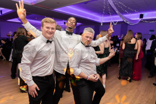 Three men dressed for a black tie evening pose for the camera. Behind, many people and dancing in the large room.