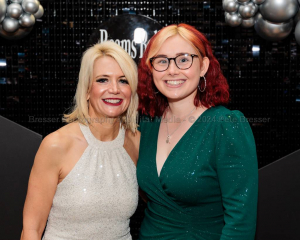 Portrait of two women, dressing in ball gowns pose for the camera in front of a glittery black backdrop with silver balloons.