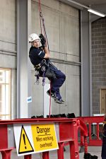 man suspended by ropes for training in building maintenance. Forwground shows 'Danger - men working overhead' sign