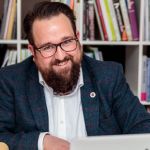 Hirsute man sitting at desk with bookcases behind him