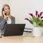 Female speaking on a mobile phone while sitting at a desk with a laptop computer and vase of flowers