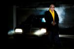businessman in dark carpark leaning on car with its lights on