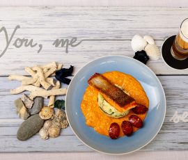 A stylised plate of cod and chorizo on a blue plate. The shot is from above and shows the plate on a rustic, limewashed wooden board. It is accompanied with a glass of beer. The board has the words 'You, me and the sea' printed on it.