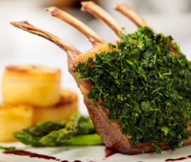 A rack of four lamb chops in close up. Behind on the plate are fondant potatoes and asparagus