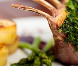 Close up of a cooked rack of lamb with a herb crust. It is accompanied by fondant potatoes and asparagus