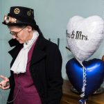 Steampunk bride checks a pocket watch. Behind here are heart shaped balloons with 'Mrs & Mrs' written on one.