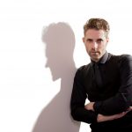 man in dark clothes leaning against a while wall with his shadow prominently shown too