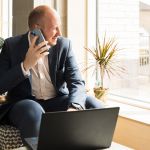 Businessman on his mobile looking out of a window. His laptop sits in front of him on a low table