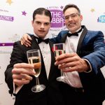 Two men, one young and dressed in 1930s style formal attire; the other, older, is in a blue velvet dress jacket and black tie. They hold champagne flute glasses in front of them. Behind them is a photo wall with many logos of corporate sponsors for the awards event