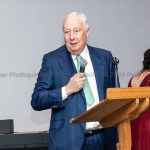 Lord Harris stands at a lecturn with a microphone in his hand. He is addressing an audience of a business awards event