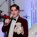 Smartly dressed young man with a small teddy bear in one hand and a microphone in the other in front of a stage. He is raising funds via an auction