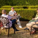 A small group of people relaxing in a sunny garden. They sit around a garden table with a few drinks to hand.